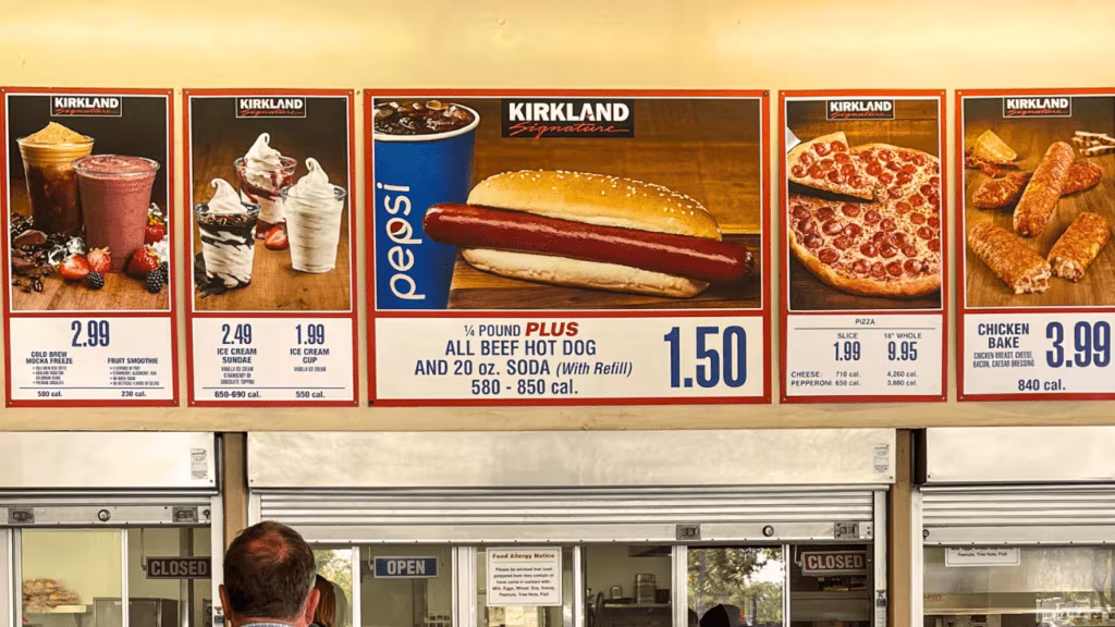 The image shows a menu board at a Costco Food Court, featuring items such as a cold brew mocha freeze, fruit smoothie, ice cream sundae or cup, all beef hot dog with soda, pizza by the slice or whole, and a chicken bake, with prices ranging from $1.50 to $9.95 and calorie information included for each item